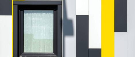 A window with a black frame and a white curtain, set in a wall with alternating yellow and white panels.