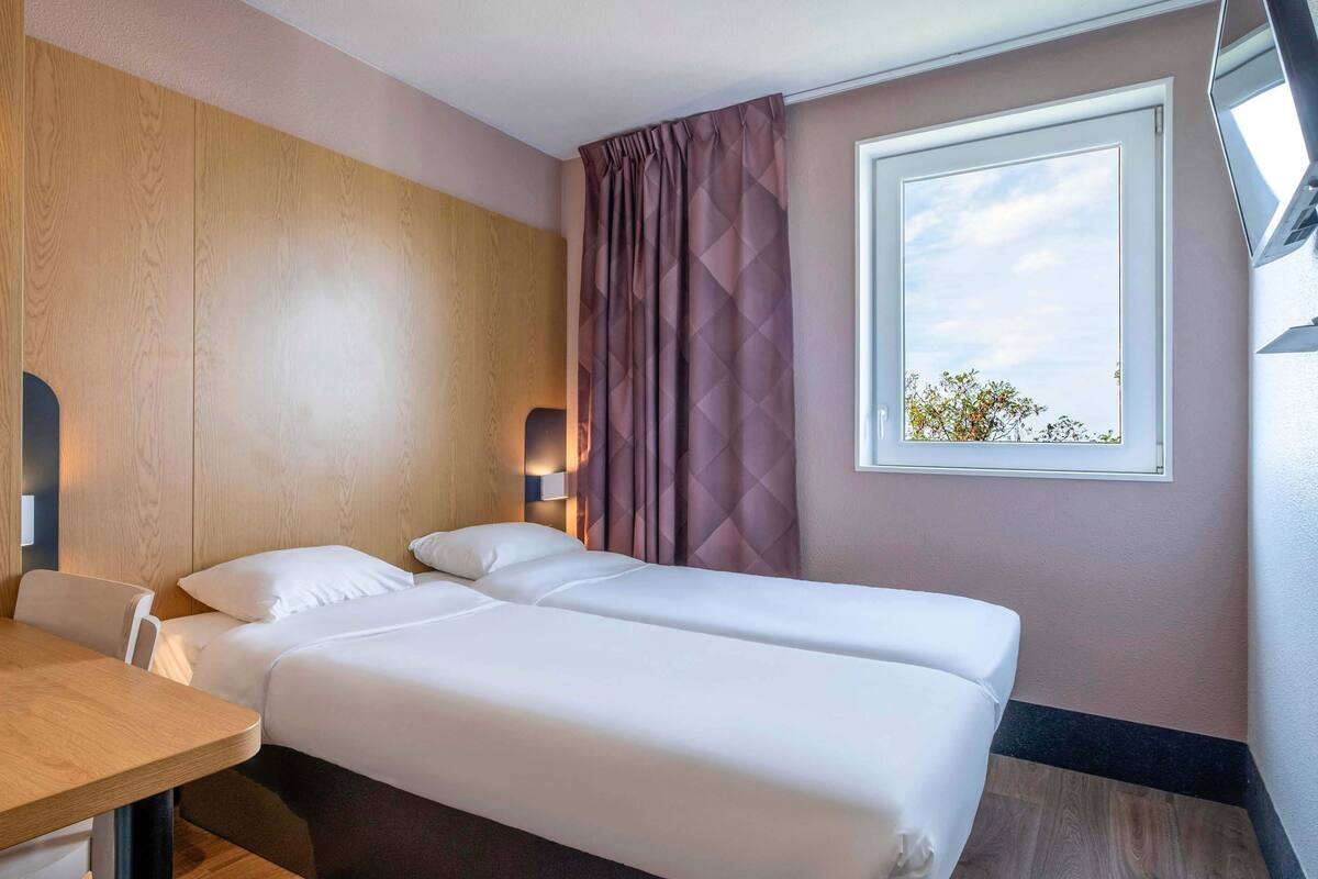 A hotel room with two beds, a wooden headboard, a desk, a chair, a window with a view, and a wall-mounted lamp.