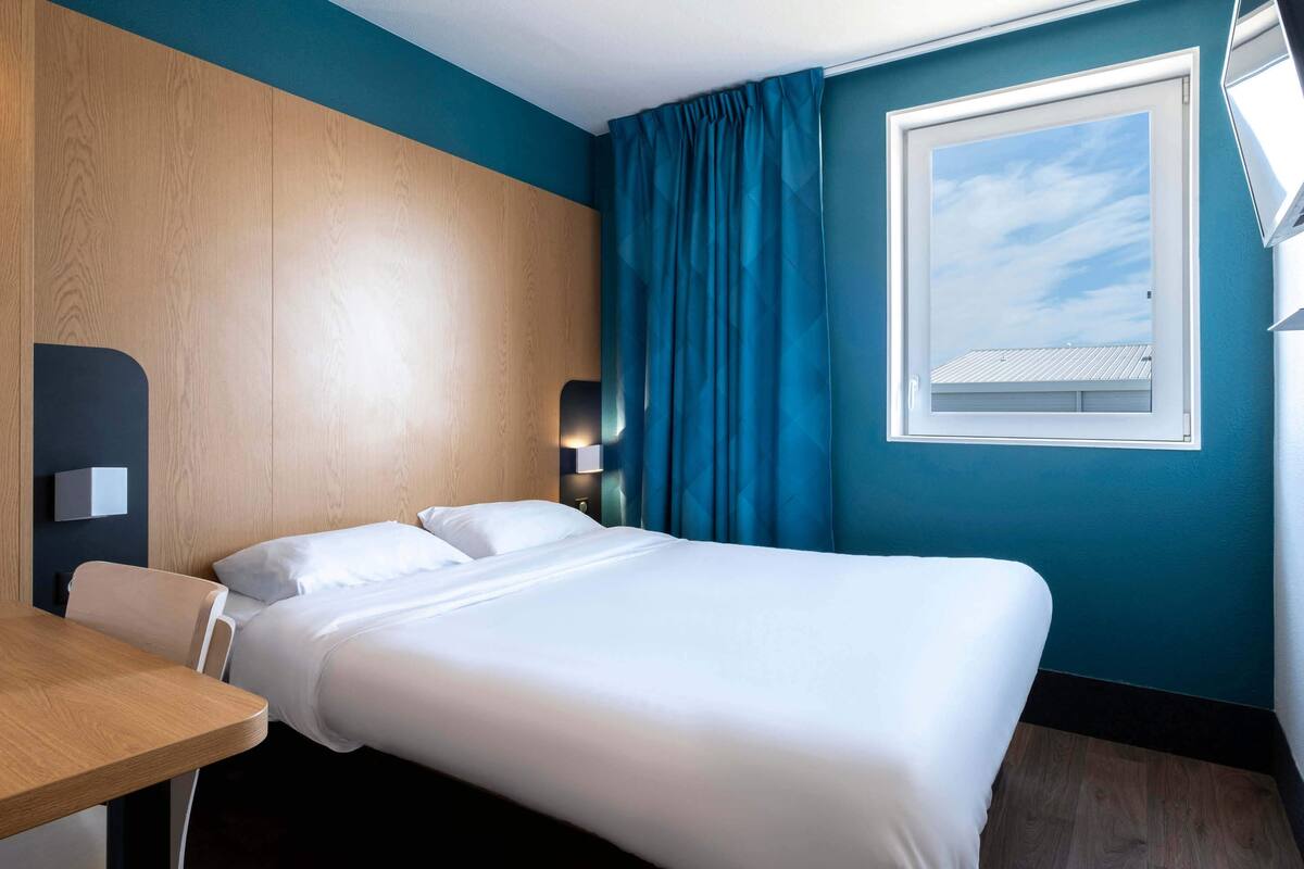 A hotel room with a bed, a desk, a window with curtains, and a view of the sky.
