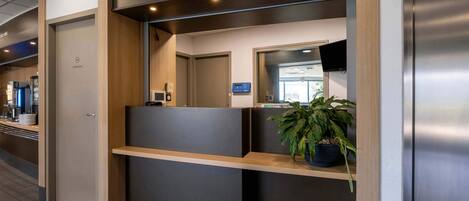 Una zona de recepción moderna con un mostrador de madera, una planta en maceta y una pantalla digital.