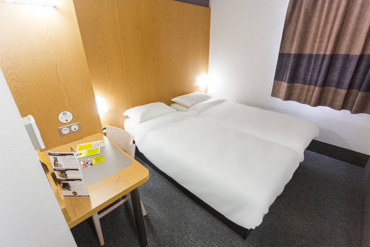 A hotel room with a wooden headboard, a desk with a chair, a bed with white bedding, and a nightstand with a lamp.