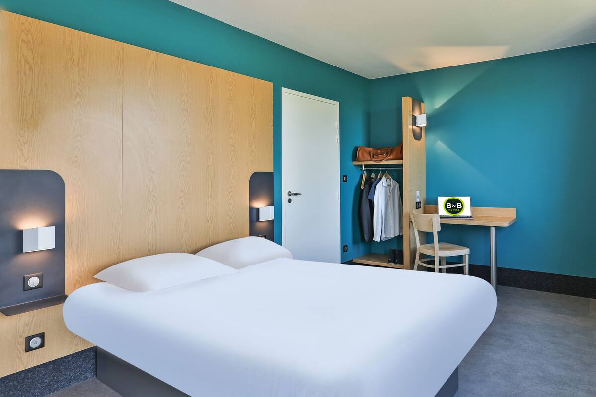 Ein modernes Hotelzimmer mit einem großen Bett, einem Schreibtisch mit Stuhl, einem Kleiderständer mit Kleidung und einer Tür.