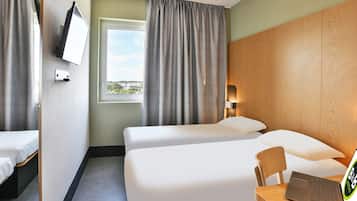 Een hotelkamer met twee bedden, een bureau met een laptop, een aan de muur bevestigde televisie en een raam met gordijnen.