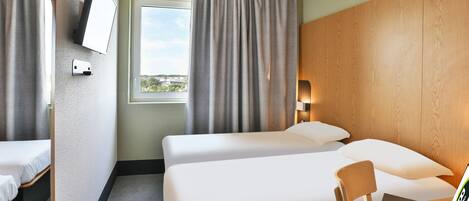 A hotel room with two beds, a desk with a laptop, a TV mounted on the wall, and a window with curtains.