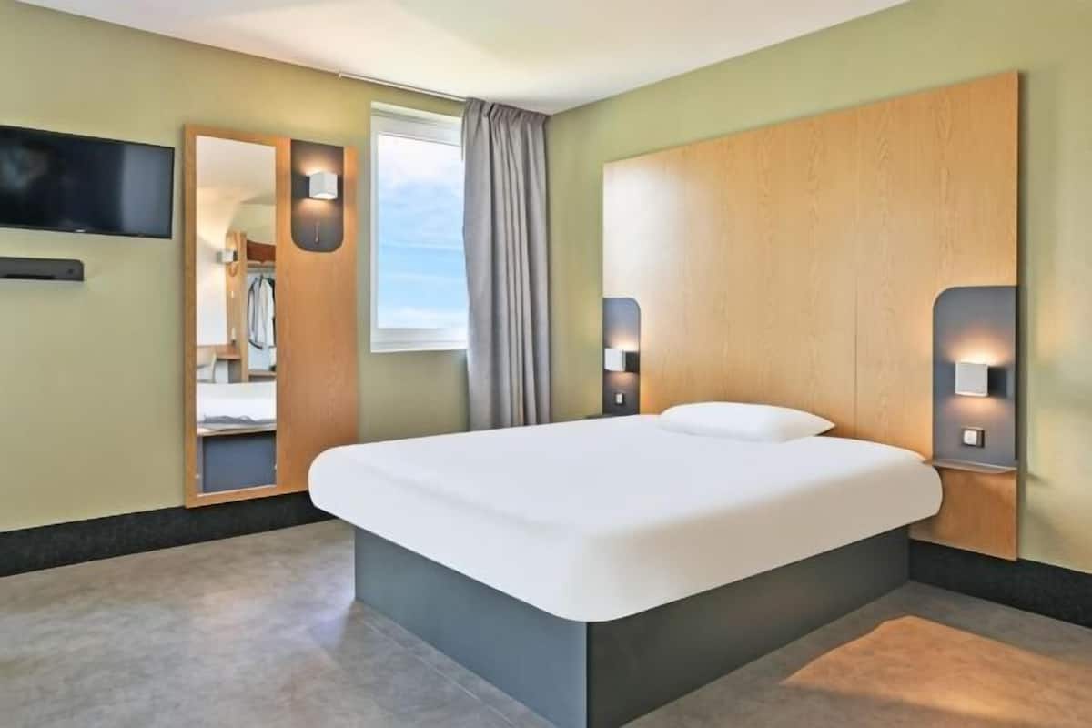 Ein Hotelzimmer mit einem großen Bett, einem an der Wand montierten Fernseher und einem Fenster mit Vorhängen.