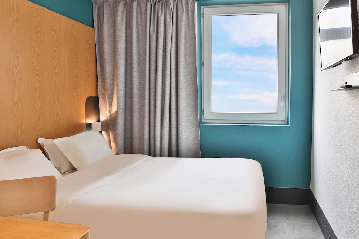 Ein Hotelzimmer mit Bett, Holz-Kopfteil, Fenster mit Vorhängen, Flachbildfernseher und Tisch.