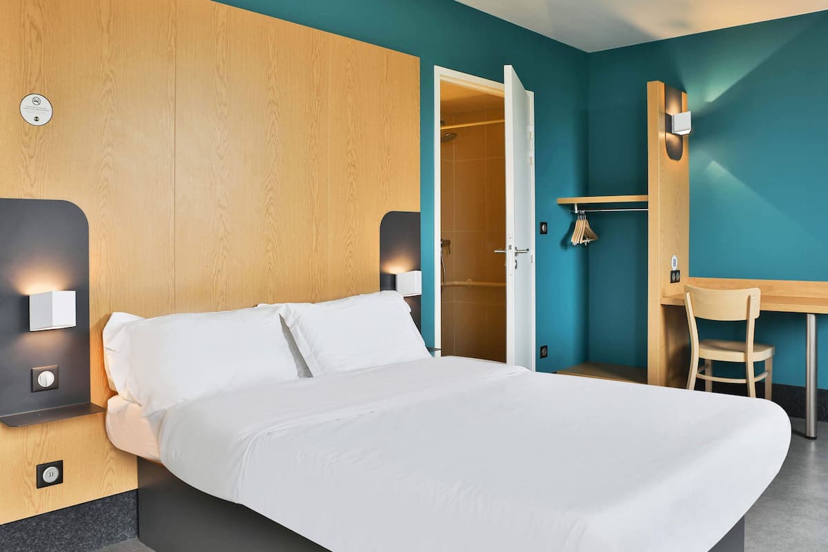 Ein Hotelzimmer mit einem Bett, einem Schreibtisch und einem Stuhl. An der Wand befindet sich ein Holzpaneel und eine Tür zu einem Badezimmer.