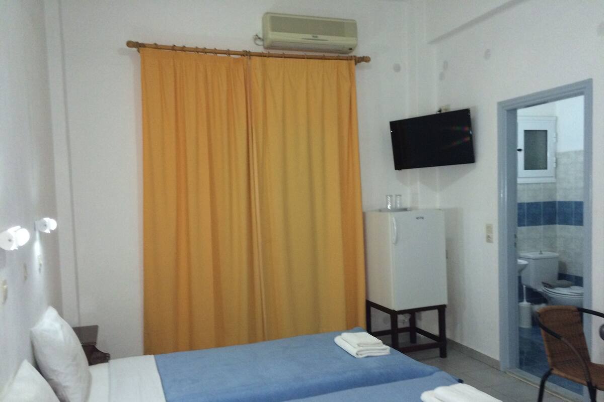 Una habitación de hotel con cama, televisor, equipo de aire acondicionado y baño visible a través de una puerta abierta.