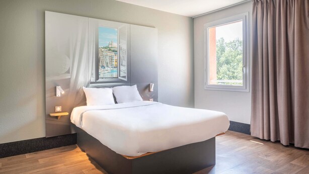 A modern hotel room with a large bed, a nightstand, a wall-mounted lamp, and a window with curtains. - B&B HOTEL Marseille Aéroport Saint-Victoret (Saint-Victoret)
