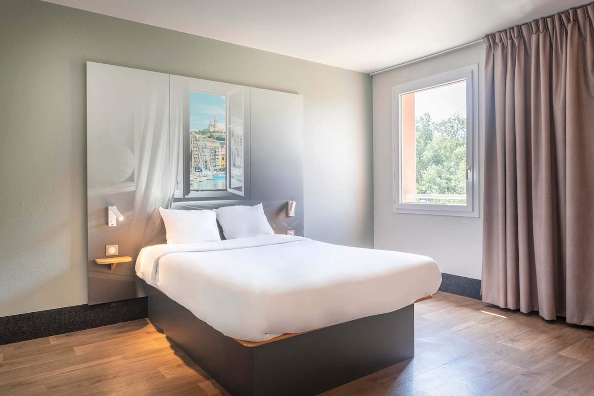 A modern hotel room with a large bed, a nightstand, a wall-mounted lamp, and a window with curtains.