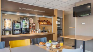 A breakfast area with a coffee machine, refrigerator, and food display. There are chairs and a table with breakfast items. - B&B HOTEL Marseille Aéroport Saint-Victoret (Saint-Victoret)