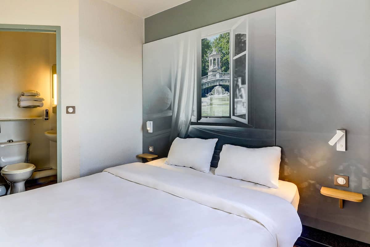 Une chambre d’hôtel avec un lit, une salle de bain dotée de toilettes et d’un lavabo, et un mur avec une grande fenêtre et un tableau.