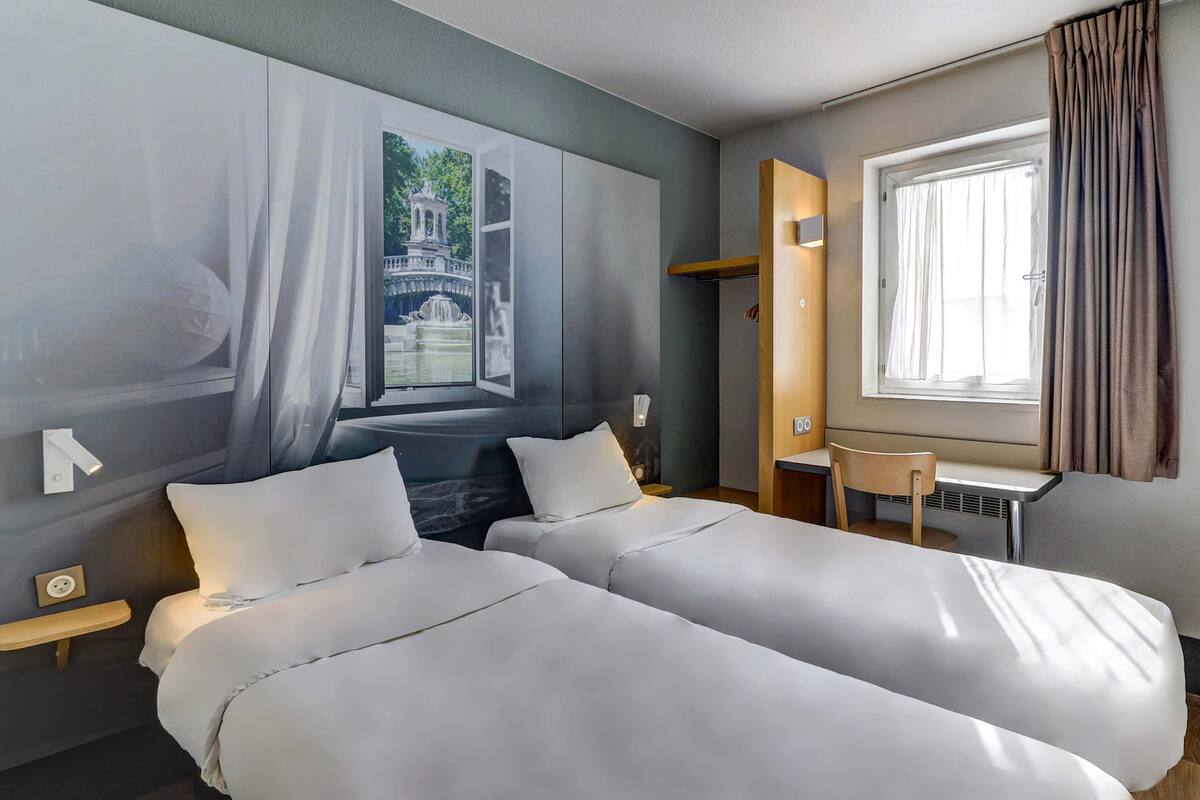 Une chambre d’hôtel avec deux lits, un petit bureau, une fenêtre avec des rideaux et un lampadaire fixé au mur.