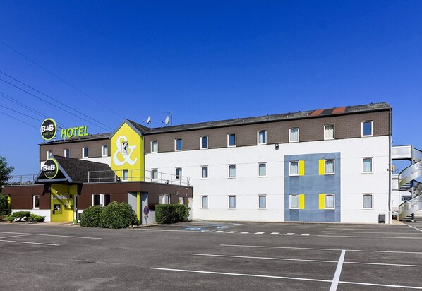 Un hôtel de la chaîne B&B, avec une façade jaune et blanche, de nombreuses fenêtres et un parking situé devant l’établissement.