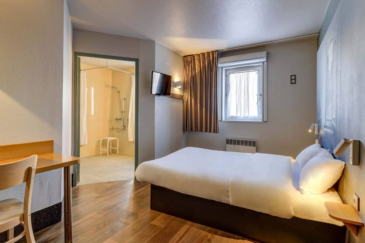 Une chambre d’hôtel avec un lit, un bureau, une chaise, une télévision et une fenêtre avec des rideaux.