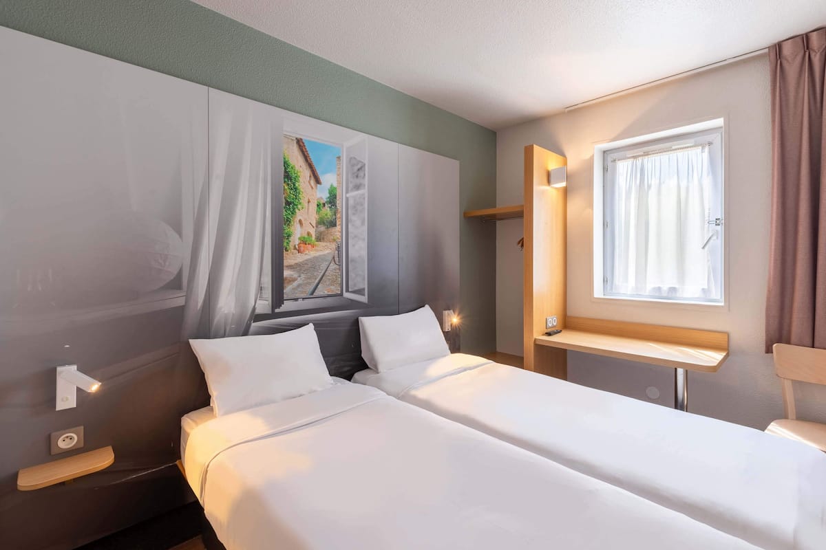 Habitación de hotel con dos camas, una ventana con cortinas, una mesita de noche y una lámpara de pared.