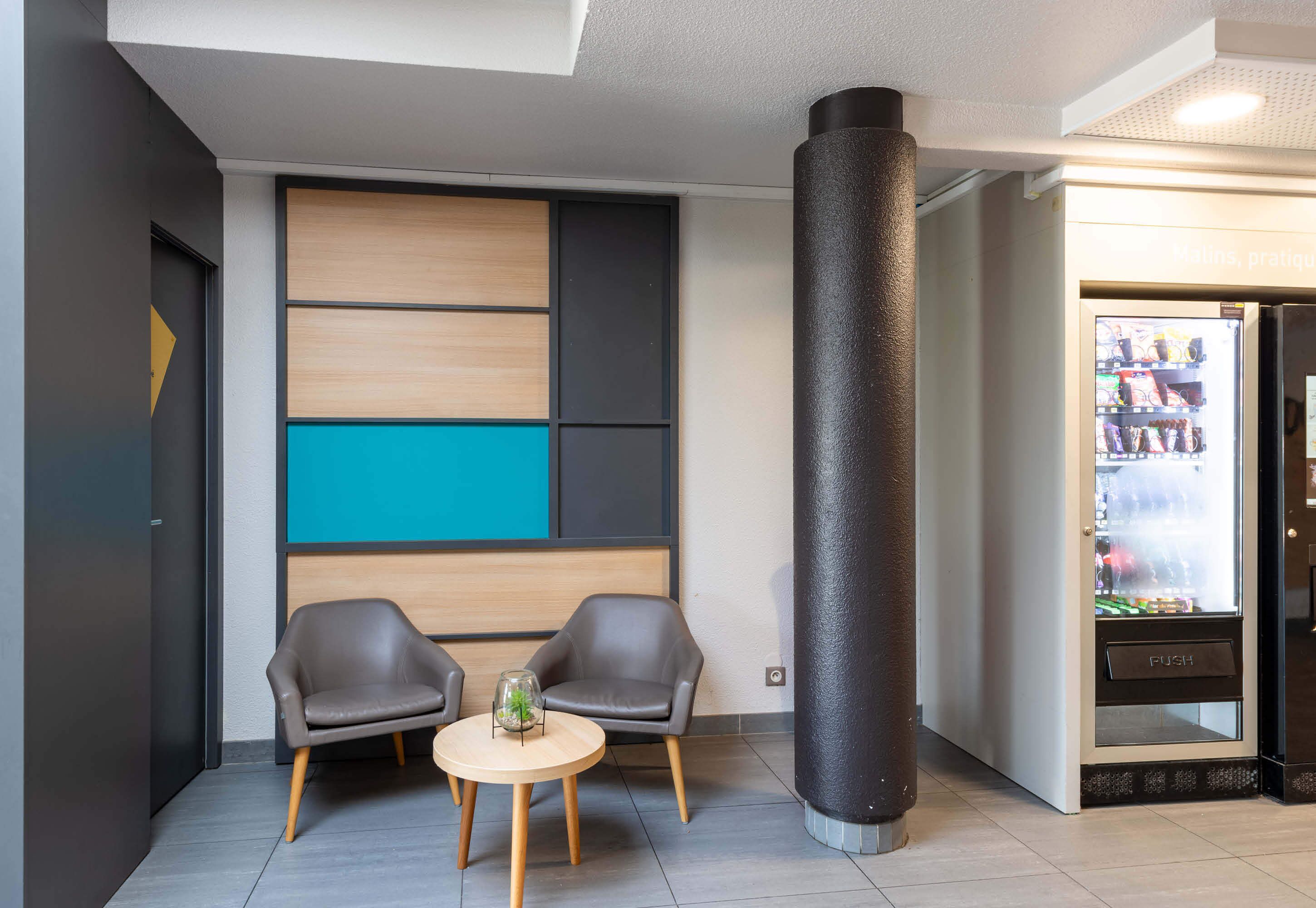 Un lobby moderno con dos sillones de cuero, una mesa redonda de madera, un pilar negro grande y una máquina expendedora.