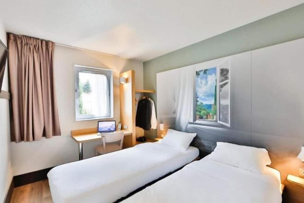 Habitación de hotel con dos camas, un escritorio, una computadora y un ventanal con cortinas.