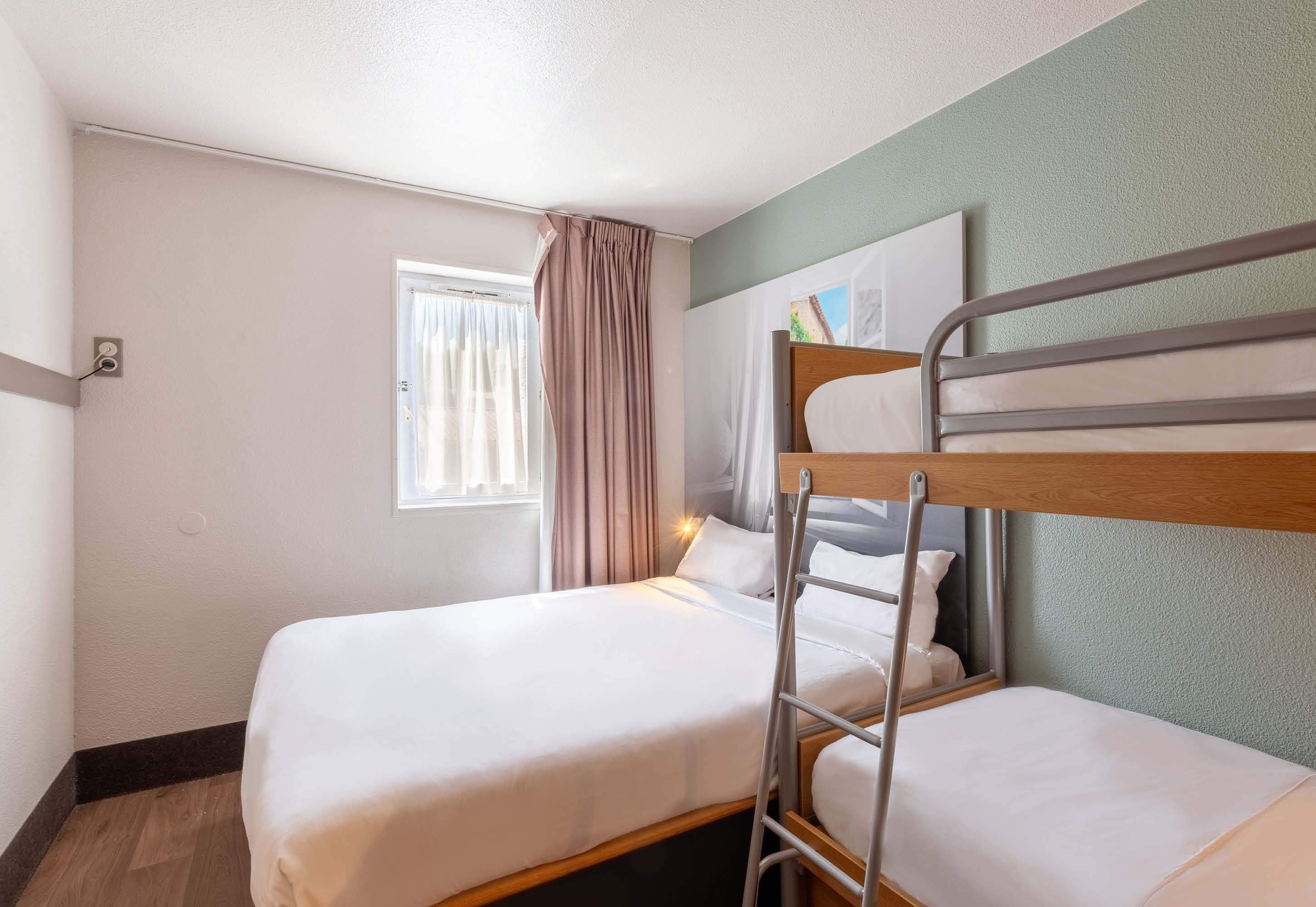Ein Zimmer mit zwei Etagenbetten, einem Vorhangfenster, einer Nachttischlampe und einem an der Wand befestigten Spiegel.