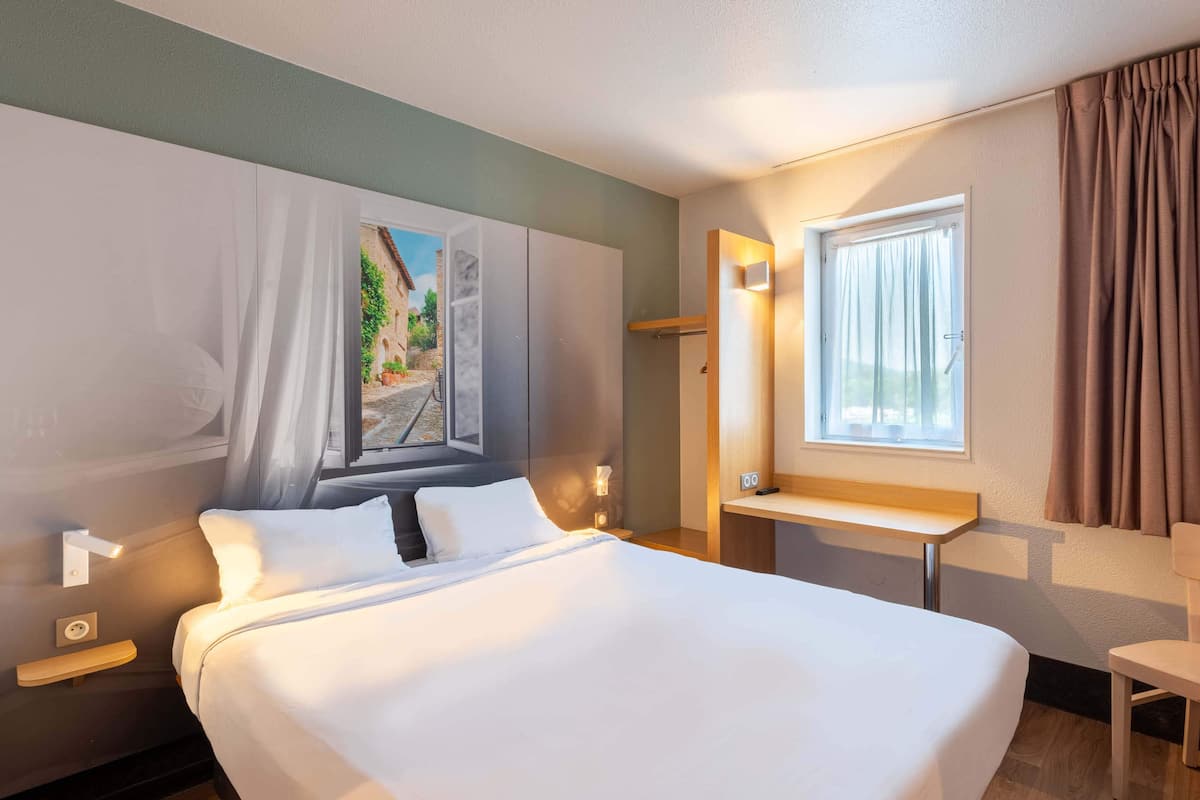 Una habitación de hotel con una cama grande, un escritorio, una ventana con cortinas y un cuadro en la pared.
