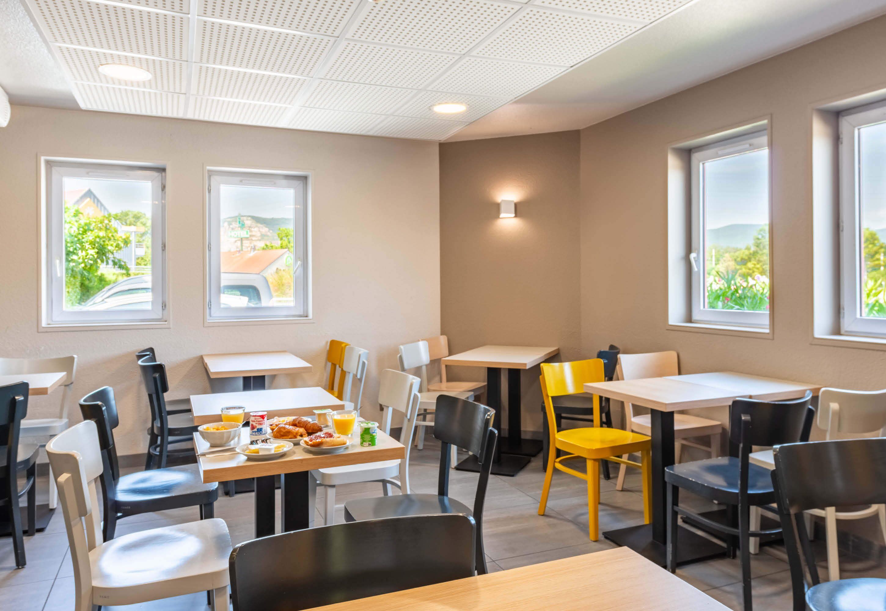 Un comedor moderno con varias mesas y sillas, ventanas amplias con vistas a zonas verdes y un techo con diseño de cuadrícula.