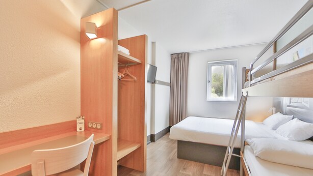 Una habitación de hotel compacta con literas, un escritorio con silla, un armario y una ventana con cortinas.