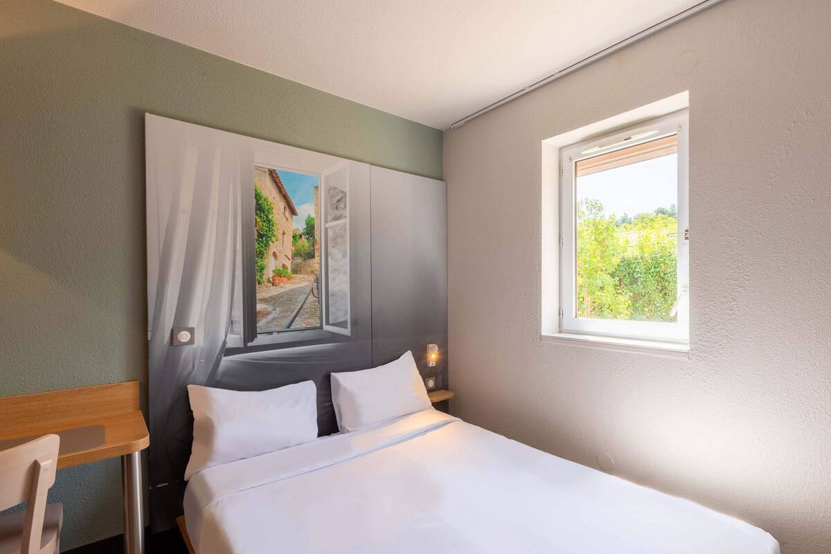 Una habitación de hotel con cama, mesita de noche, ventana y un cuadro en la pared.