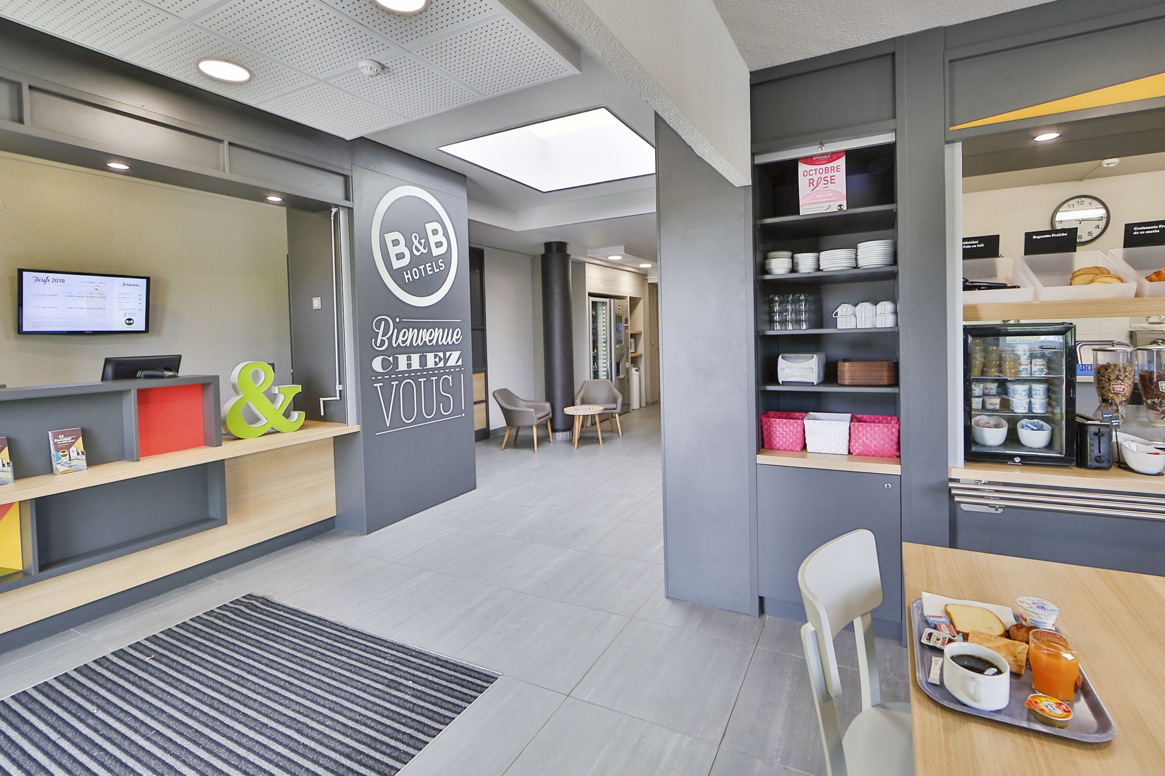 A B&B hotel reception area with a digital display, a reception desk, and a breakfast area with a tray of food and beverages.