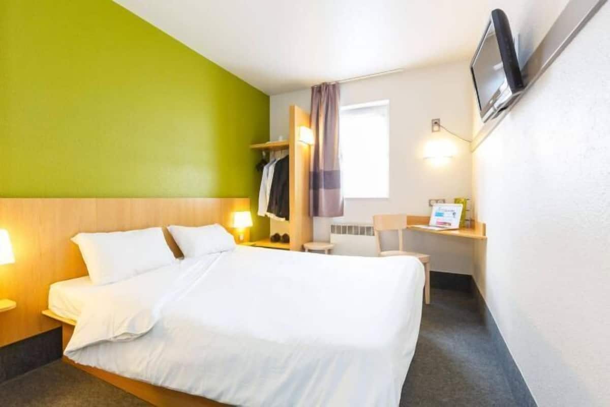 Una habitación de hotel con cama, mesita de noche, lámpara, televisor y un pequeño escritorio con una silla.