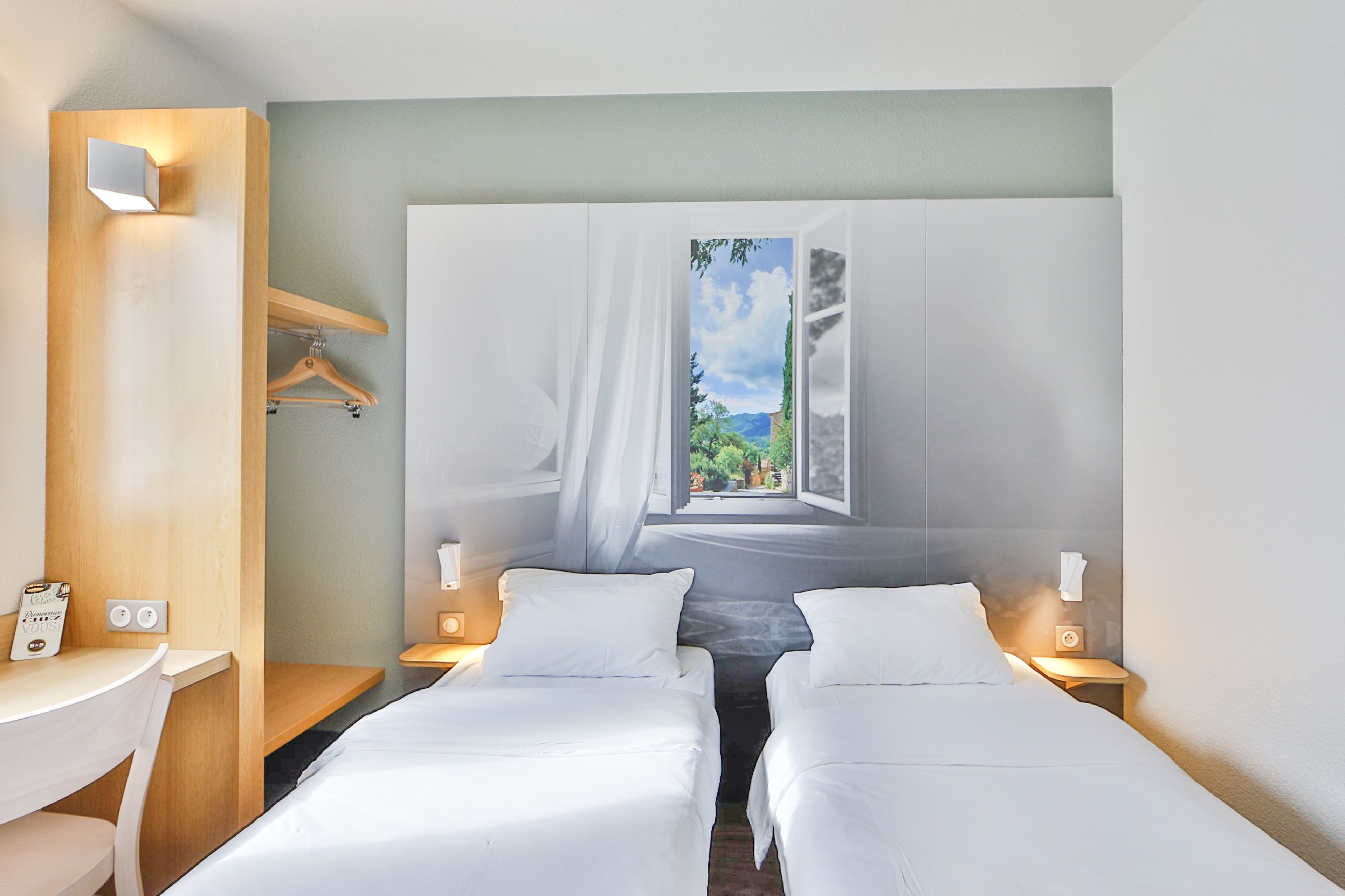 Een hotelkamer met twee bedden, een bureau en een groot raam met uitzicht op groen.