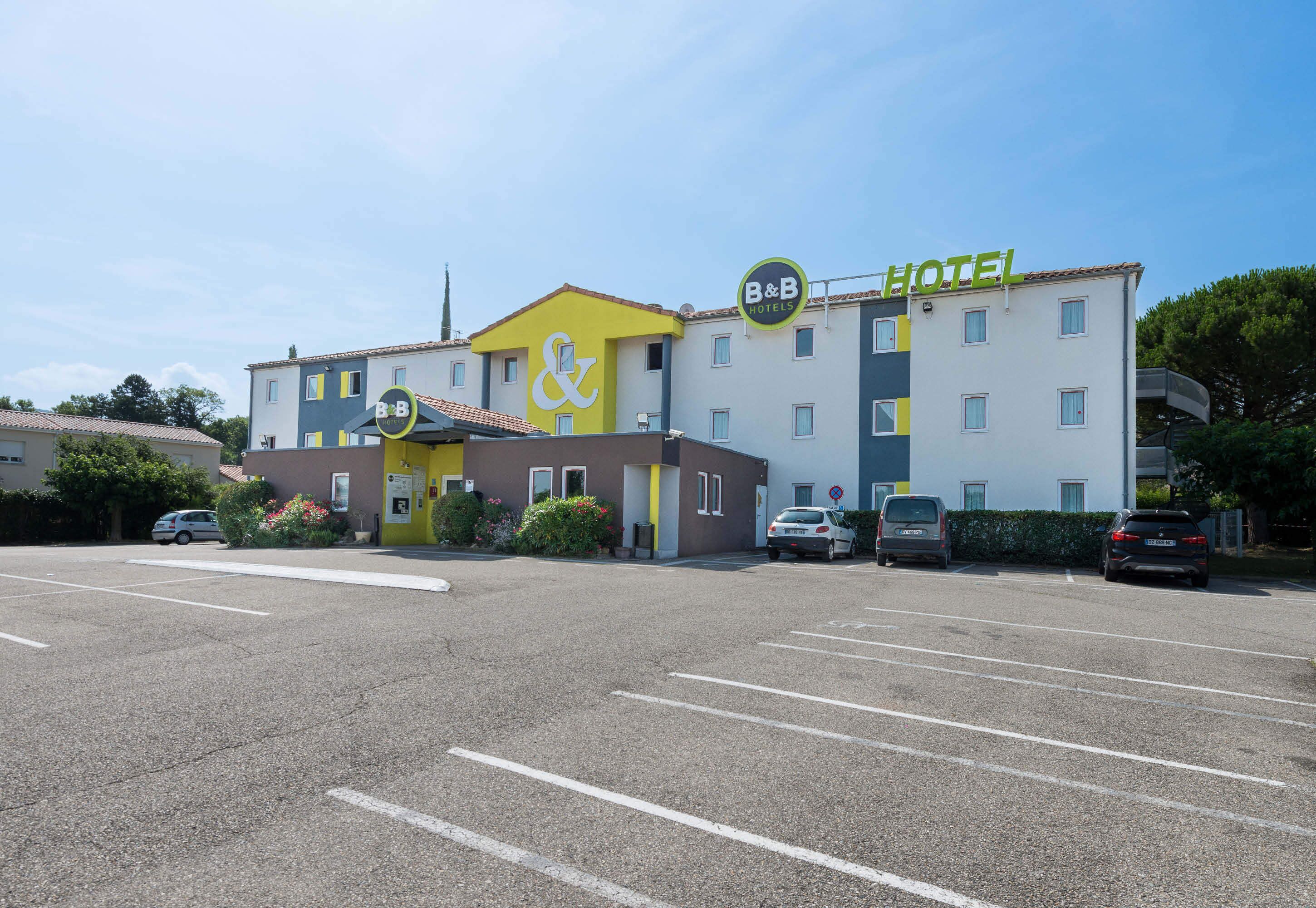 A B&B hotel with a yellow and white facade, multiple parking spaces, and a clear blue sky.