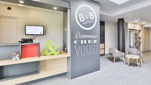 Un área de recepción de hotel con diseño moderno, que incluye un mostrador de recepción, una pantalla digital y un cartel de bienvenida.