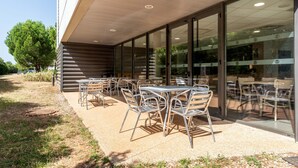 Un café en plein air avec des tables et des chaises, un bâtiment doté de grandes fenêtres et un arbre en arrière-plan.
