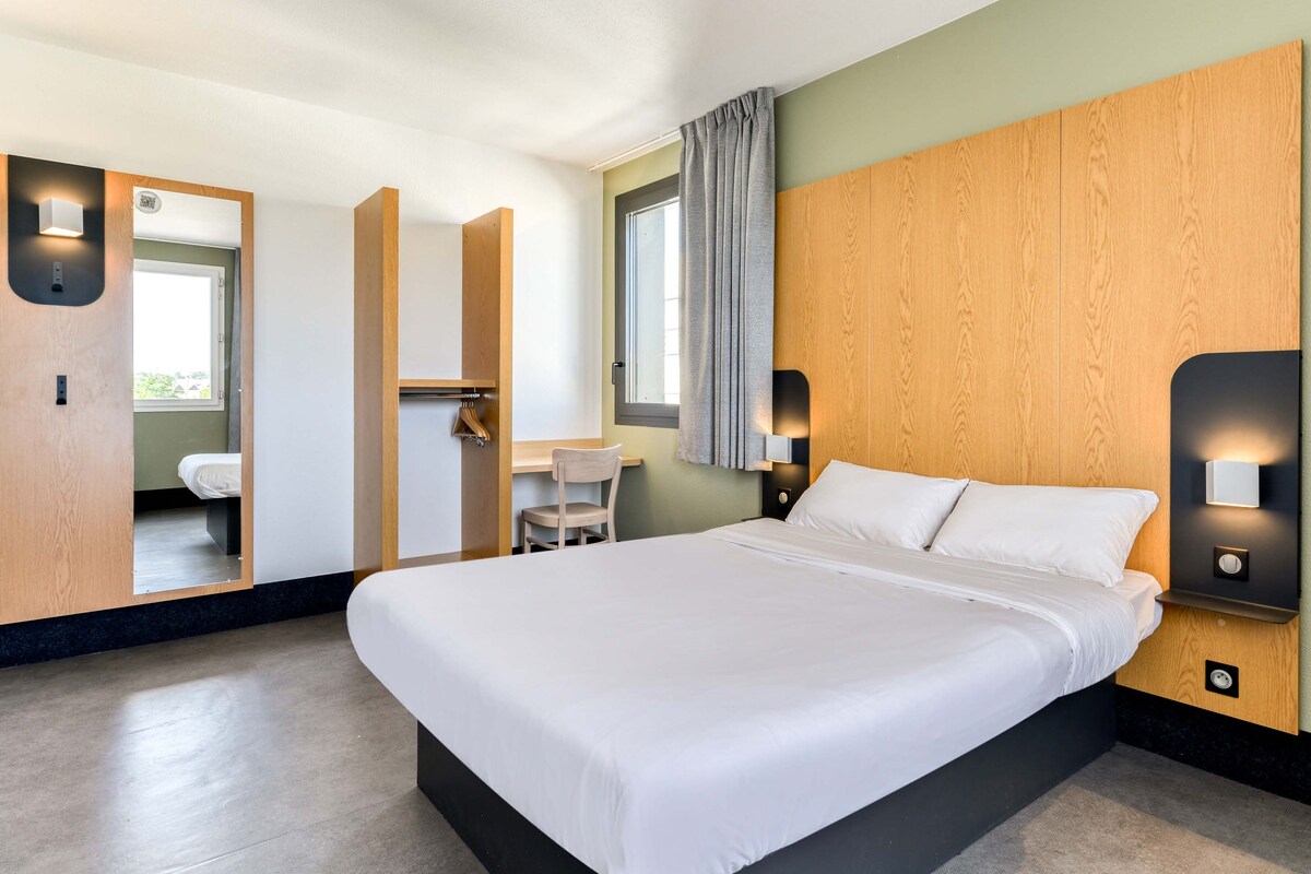 Ett modernt hotellrum med en stor säng, ett skrivbord och en stol. Det finns ett fönster med gardiner och utsikt över omgivningen.