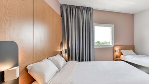 Une chambre d’hôtel avec une tête de lit en bois, un grand lit, un lit plus petit, une fenêtre avec des rideaux et des lampes de chevet.