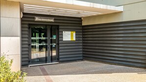 Une entrée d'immeuble avec une pancarte « Bienvenue », une porte vitrée et, à droite, un volet métallique fermé.