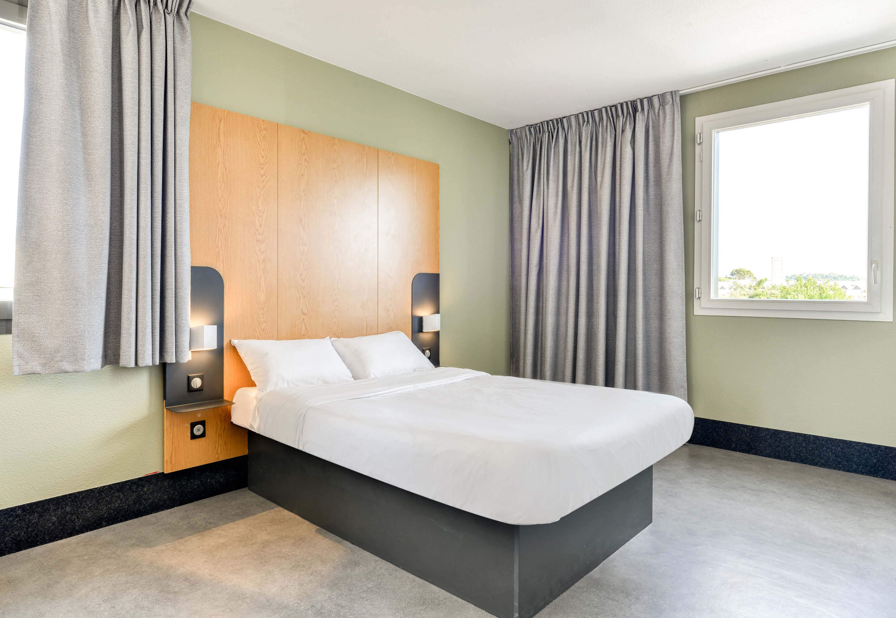 Ein modernes Hotelzimmer mit einem großen Bett, einem Holzkopfende, zwei Nachttischen inklusive Lampen sowie einem Fenster mit Vorhängen.