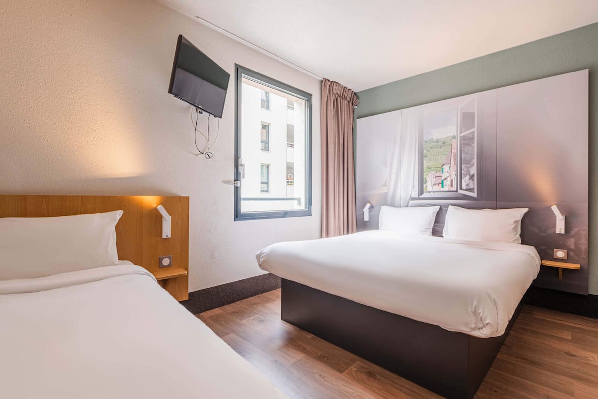 A hotel room with two beds, a TV mounted on the wall, a window with curtains, and a view of a building outside.