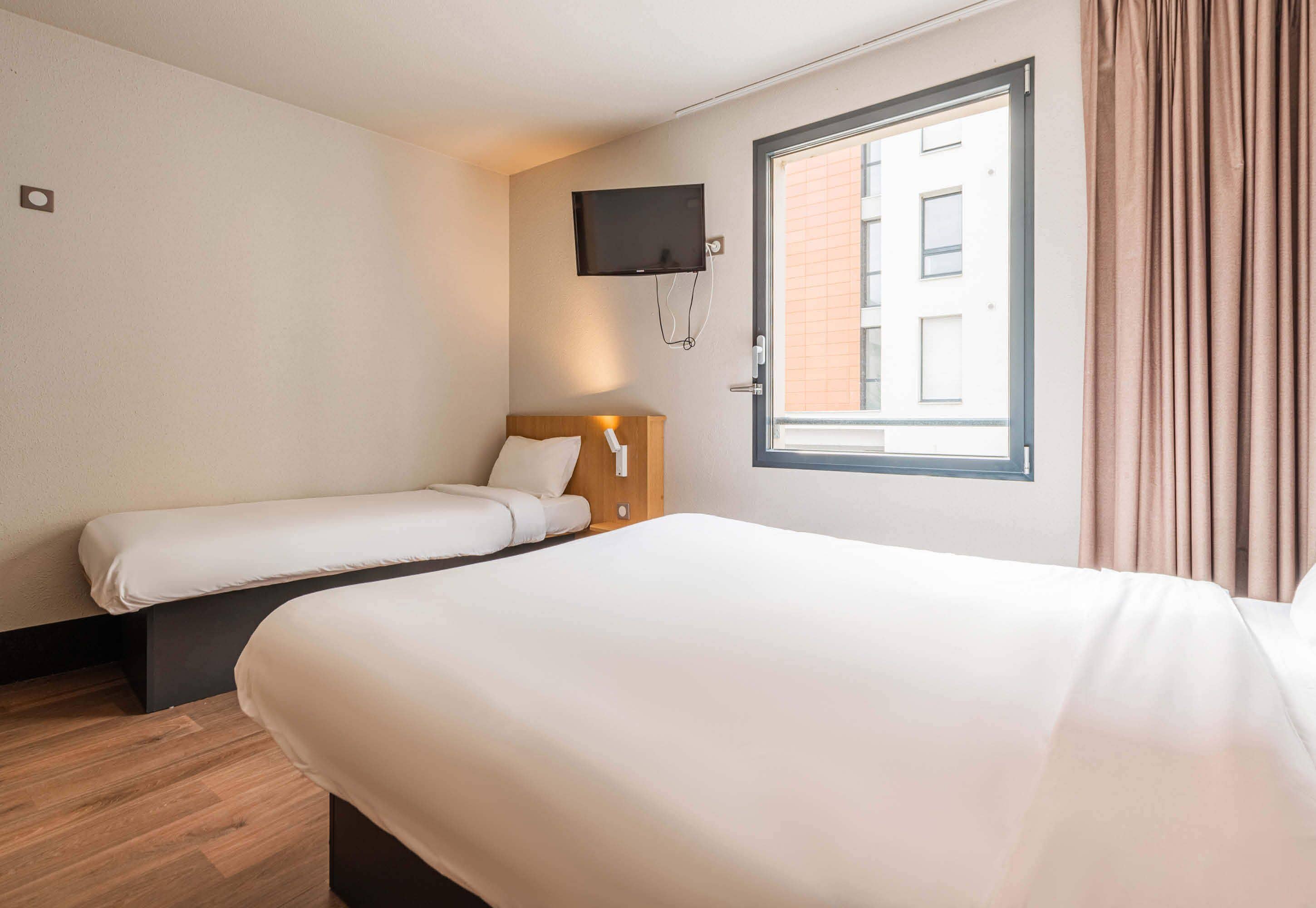 Quarto de hotel com duas camas, televisão fixada na parede e janela com cortinas.