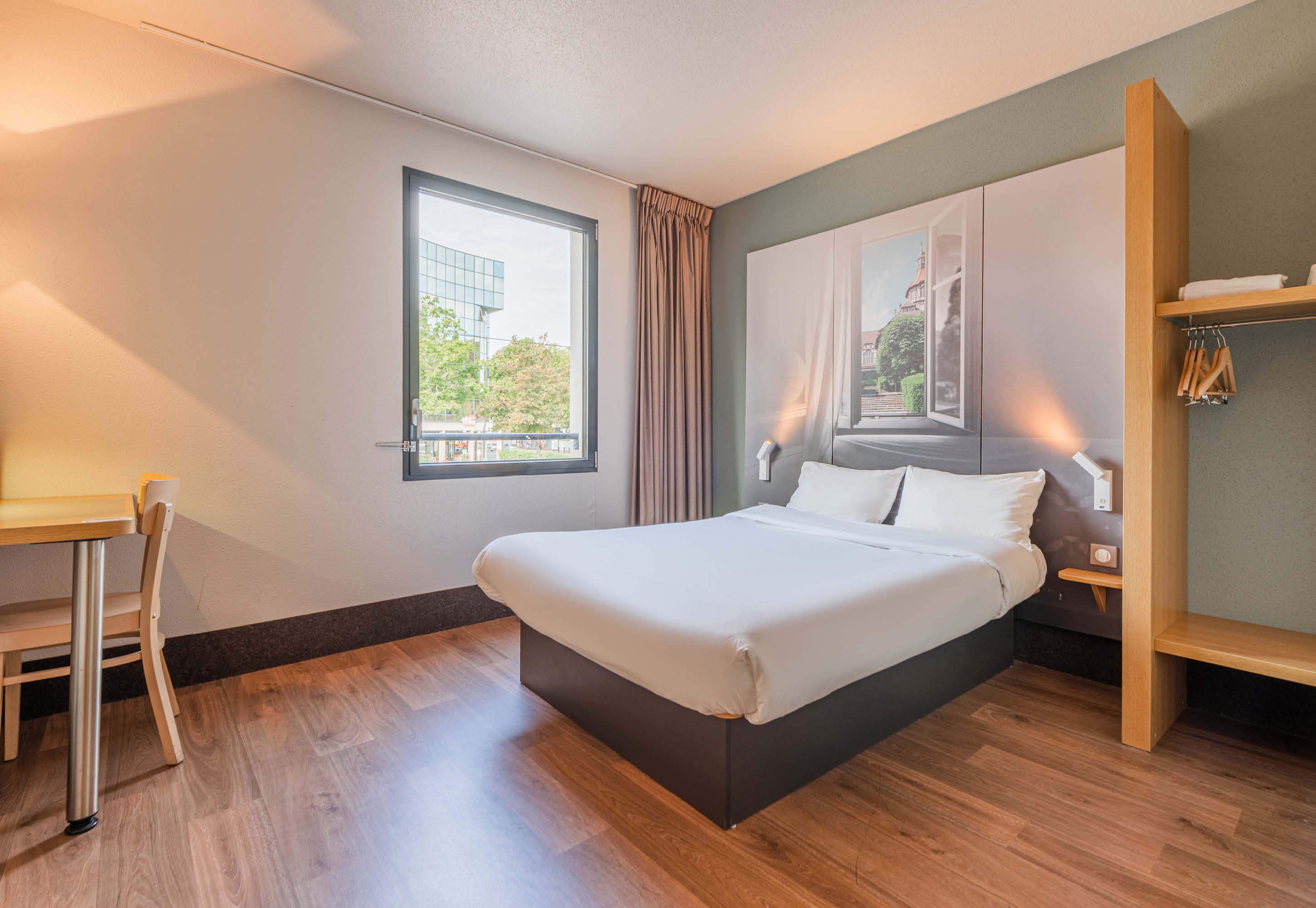 A modern hotel room with a large bed, a desk, a window with curtains, and a view of a building outside.