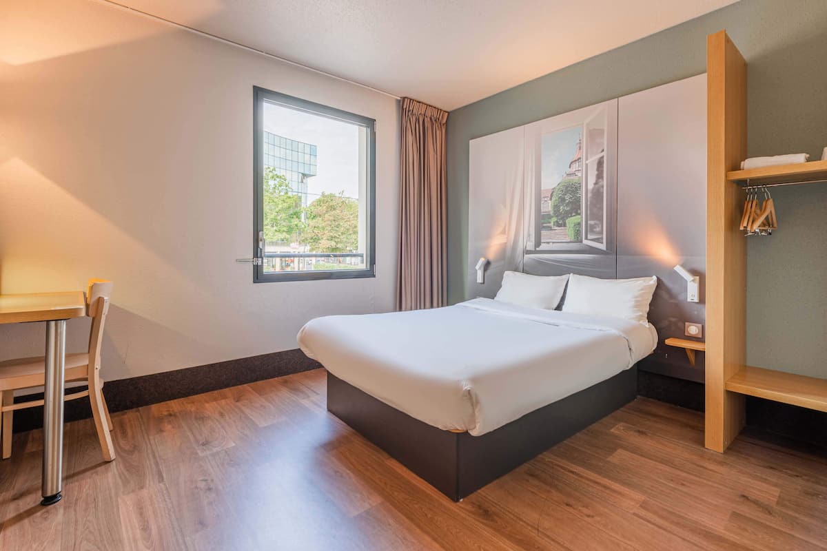 A modern hotel room with a large bed, a desk, and a window with a view of a building and trees.