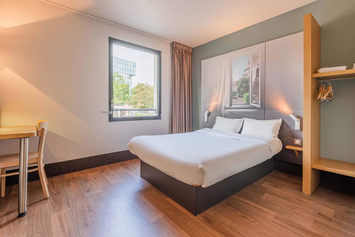 A modern hotel room with a large bed, a desk, and a window with a view of a building and trees.