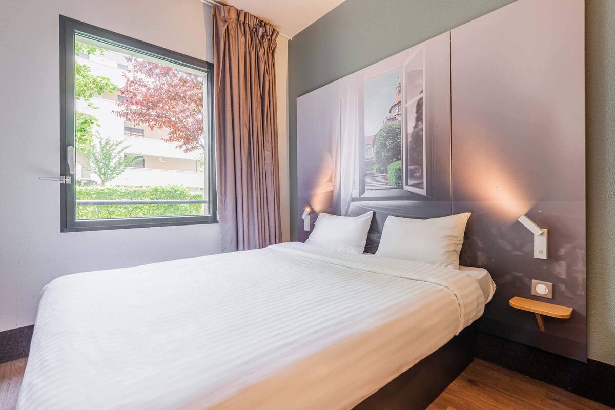 A neatly made bed with white linens, a window with curtains, and a wall-mounted nightstand with a lamp.