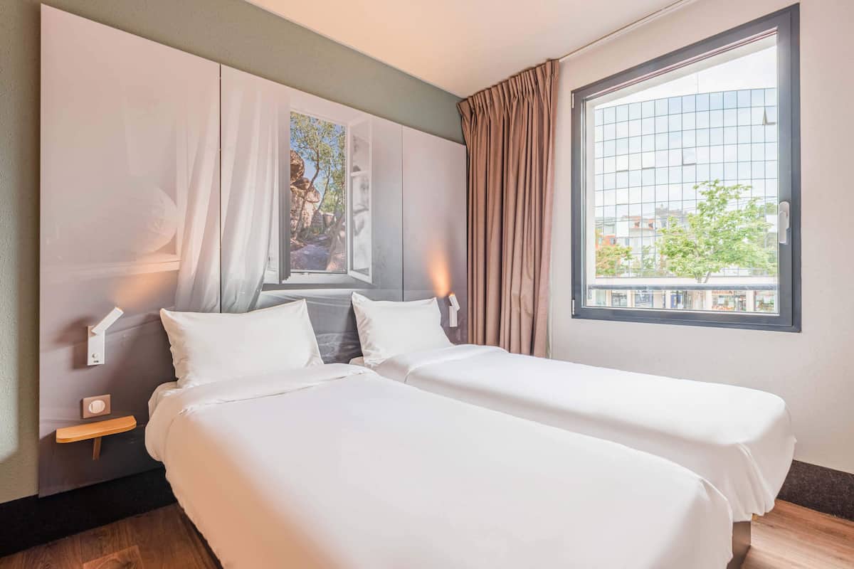 A hotel room with two beds, a large window with curtains, a painting on the wall, and a nightstand with a lamp.