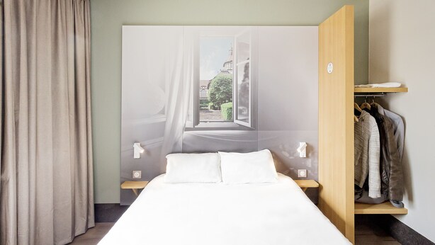 A bedroom with a bed, a nightstand, a wooden wardrobe, and a view of a building through an open window. - B&B HOTEL Mulhouse Centre (Mulhouse)