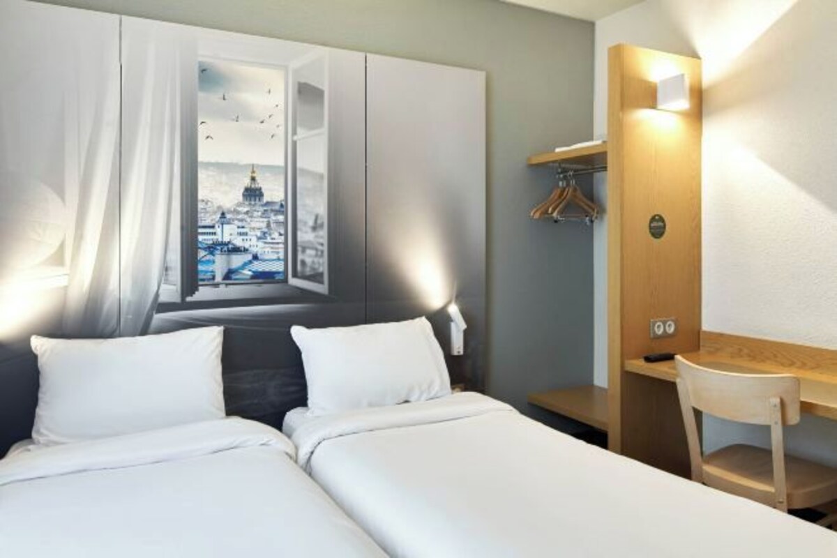 Habitación de hotel con cama, escritorio y silla. Se observa un paisaje urbano a través de una ventana con cortinas.