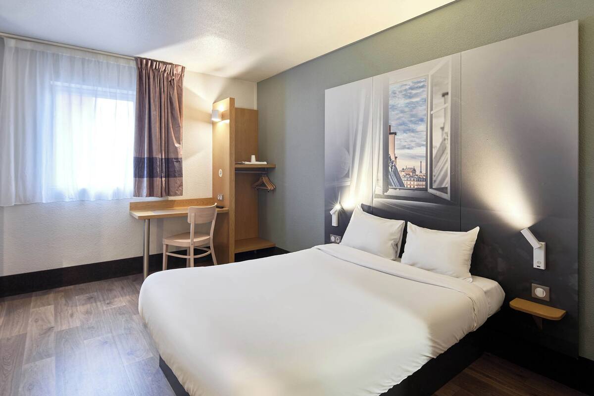 Quarto de hotel com uma cama grande, uma secretária com cadeira, um guarda-roupa e uma mesa de cabeceira com abajur.