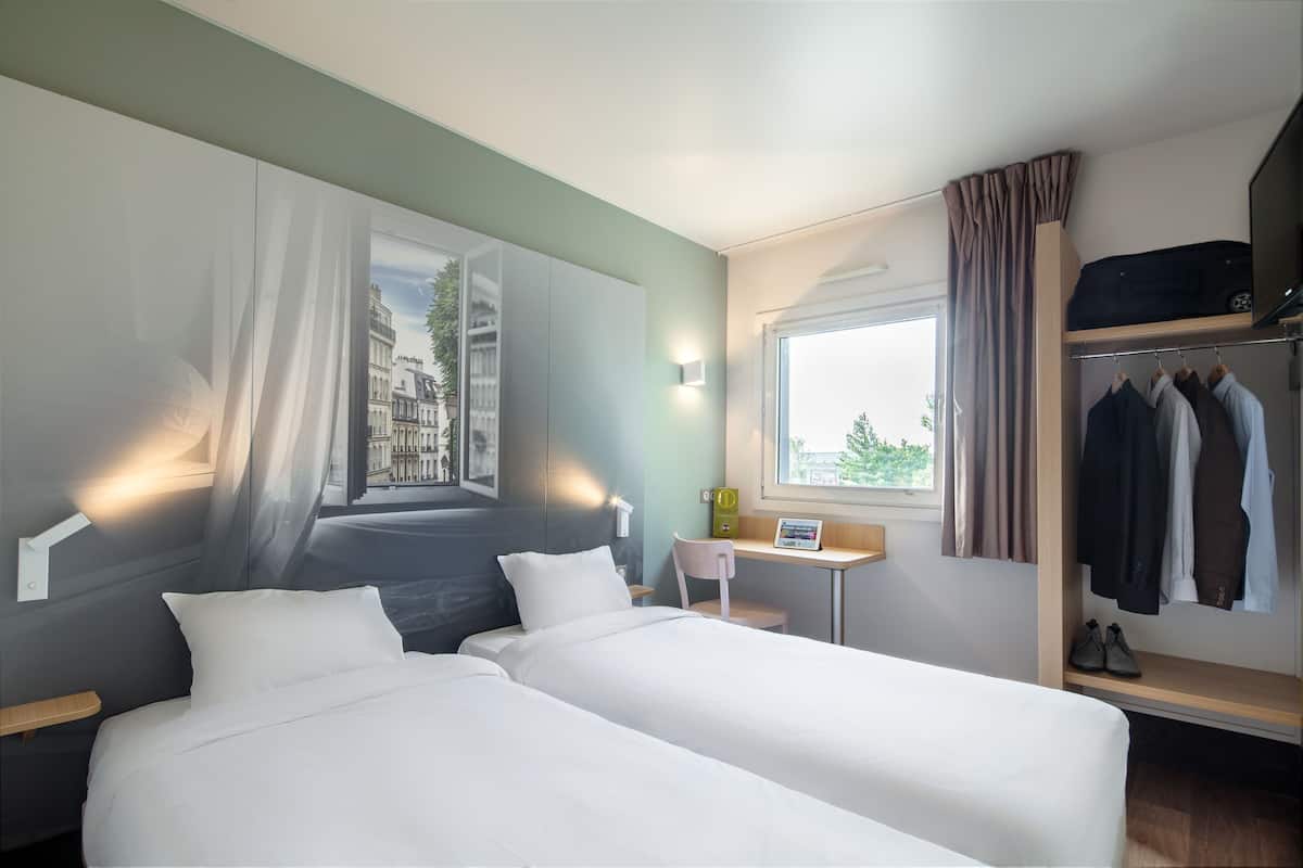 Une chambre d’hôtel avec deux lits, un bureau, une chaise, une armoire et une fenêtre avec des rideaux.