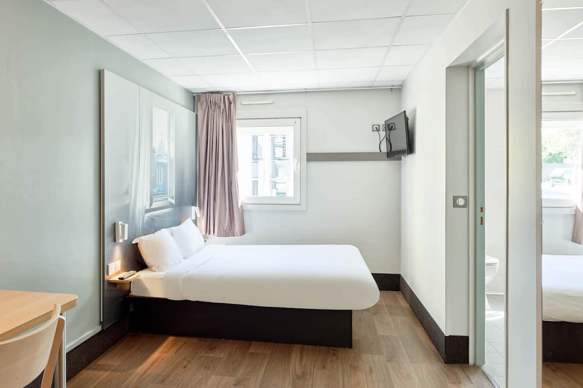 Une chambre d’hôtel moderne équipée d’un lit, d’un bureau, d’une télévision et d’une fenêtre avec des rideaux.