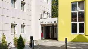 A building with a sign reading "ACCUEIL" at the entrance, a yellow building to the right, and a paved area with plants in front.
